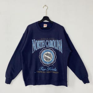 K&ouml;nnte beinhalten: Marineblaues Sweatshirt mit dem Schriftzug "NORTH CAROLINA" in einer verzierten Schriftart. Darunter befindet sich ein kreisf&ouml;rmiges Emblem mit den Worten "UNIVERSITY OF NORTH CAROLINA" und "Tar Heels". Das Sweatshirt hat lange &Auml;rmel.