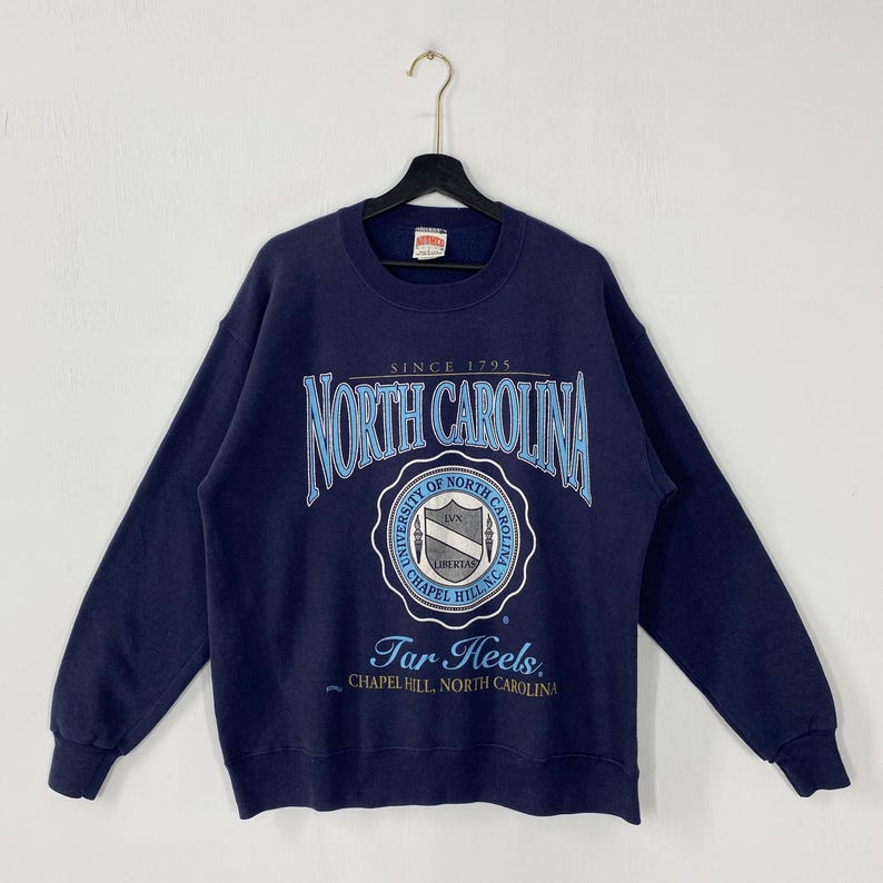 K&ouml;nnte beinhalten: Marineblaues Sweatshirt mit dem Schriftzug "NORTH CAROLINA" in einer verzierten Schriftart. Darunter befindet sich ein kreisf&ouml;rmiges Emblem mit den Worten "UNIVERSITY OF NORTH CAROLINA" und "Tar Heels". Das Sweatshirt hat lange &Auml;rmel.