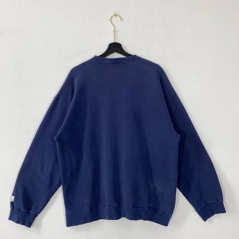May include: A navy blue sweatshirt hangs on a black hanger against a white background. The sweatshirt has long sleeves and a crew neck. The fabric appears to be a soft, slightly faded material. A small white logo is visible on the left sleeve cuff.