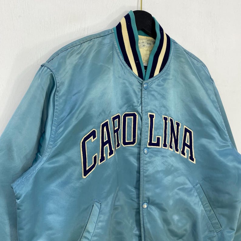 May include: Light blue satin jacket with the word "CAROLINA" embroidered across the chest in navy blue. The jacket has a striped collar in navy, teal, and cream. The jacket is buttoned up and hanging on a black hanger.