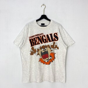 May include: A light gray heathered short-sleeve t-shirt featuring a graphic of the Tasmanian Devil character with white bandages on its hands. Above the character, the text reads CINCINNATI BENGALS in bold black letters with orange outlines.