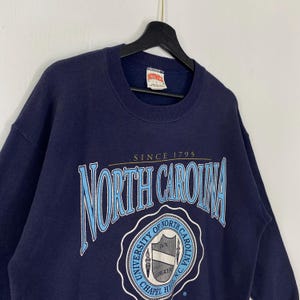 K&ouml;nnte beinhalten: Marineblaues Sweatshirt mit dem Schriftzug "NORTH CAROLINA" in einer stilisierten Schriftart. Das Sweatshirt hat auch den Text "SINCE 1795" und ein Universit&auml;tslogo. Das Sweatshirt h&auml;ngt an einem schwarzen Kleiderb&uuml;gel.