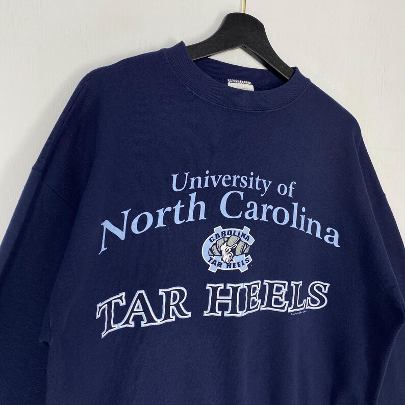 May include: Navy blue sweatshirt with the text "University of North Carolina" in light blue, and a Tar Heels logo. The sweatshirt is suspended from a black hanger.