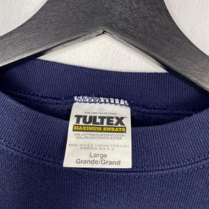 May include: Navy blue sweatshirt with a white label. The label reads "TULTEX MAXIMUM SWEATS" in black and yellow. The label also states the sweatshirt is 50% cotton and 50% polyester, and is a size Large.