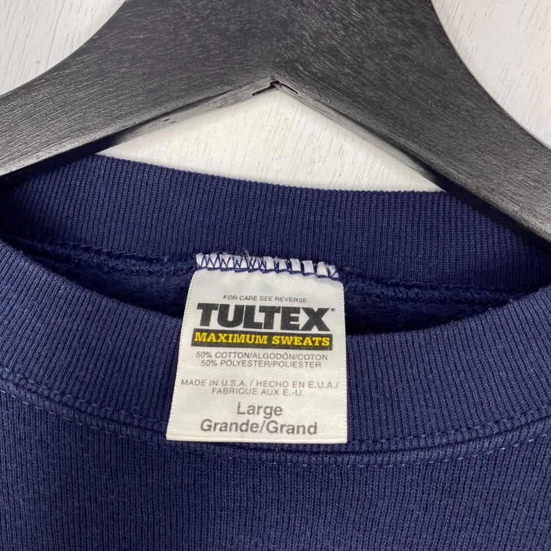 May include: Navy blue sweatshirt with a white label. The label reads "TULTEX MAXIMUM SWEATS" in black and yellow. The label also states the sweatshirt is 50% cotton and 50% polyester, and is a size Large.