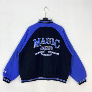 Puede incluir: Una chaqueta universitaria negra y azul con el logotipo de Orlando Magic bordado en la espalda. El texto dice MAGIC, Orlando Magic y National Basketball Association. Tiene cuello, puños y dobladillo acanalados con rayas azules y blancas.