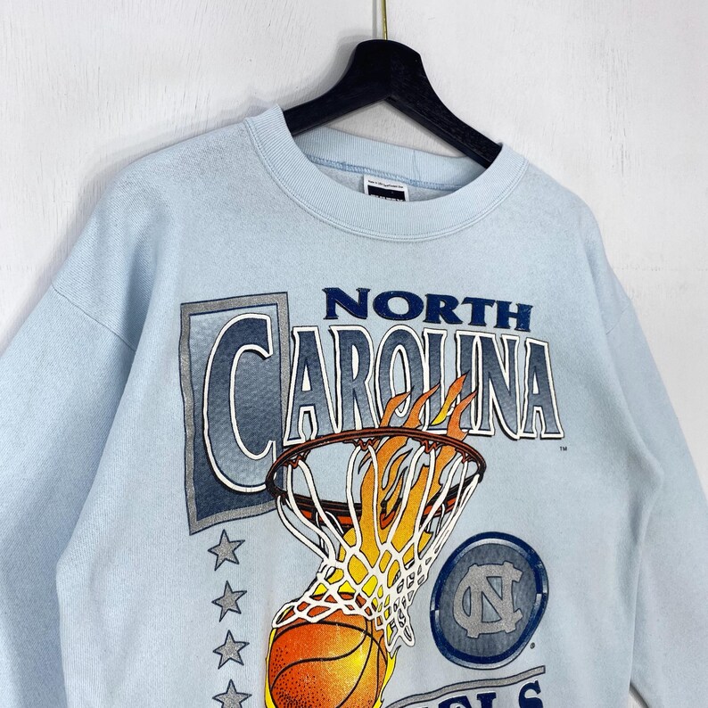 May include: A light blue crewneck sweatshirt featuring a graphic of a basketball going through a net with flames. The text reads NORTH CAROLINA, with a column of stars on the left and a circular logo containing the letters NC on the right side.
