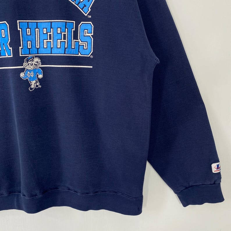 May include: Navy blue sweatshirt with the text "R HEELS" in white and light blue block letters. A cartoon Tar Heel mascot is below the text. The sweatshirt has a crew neck and long sleeves.