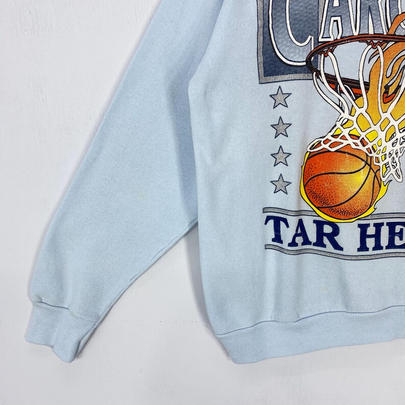 May include: A light blue long-sleeve sweatshirt featuring a graphic of a basketball going through a hoop with flames. The design includes four grey stars and the text "CAROLINA" and "TAR HEELS" in dark blue lettering on the front of the garment.