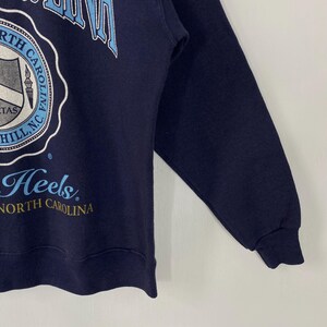 K&ouml;nnte beinhalten: Marineblaues Sweatshirt mit den W&ouml;rtern "North Carolina" in Hellblau und Wei&szlig;. Das Sweatshirt hat ein rundes Emblem mit Text und einer Grafik. Das Wort "Heels" ist ebenfalls sichtbar.