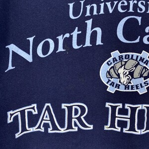 May include: Navy blue sweatshirt with the words "North Carolina" in light blue, and a "Tar Heels" logo. The logo features a ram's head in a light blue and grey colour scheme. The words "TAR HEELS" are also visible.