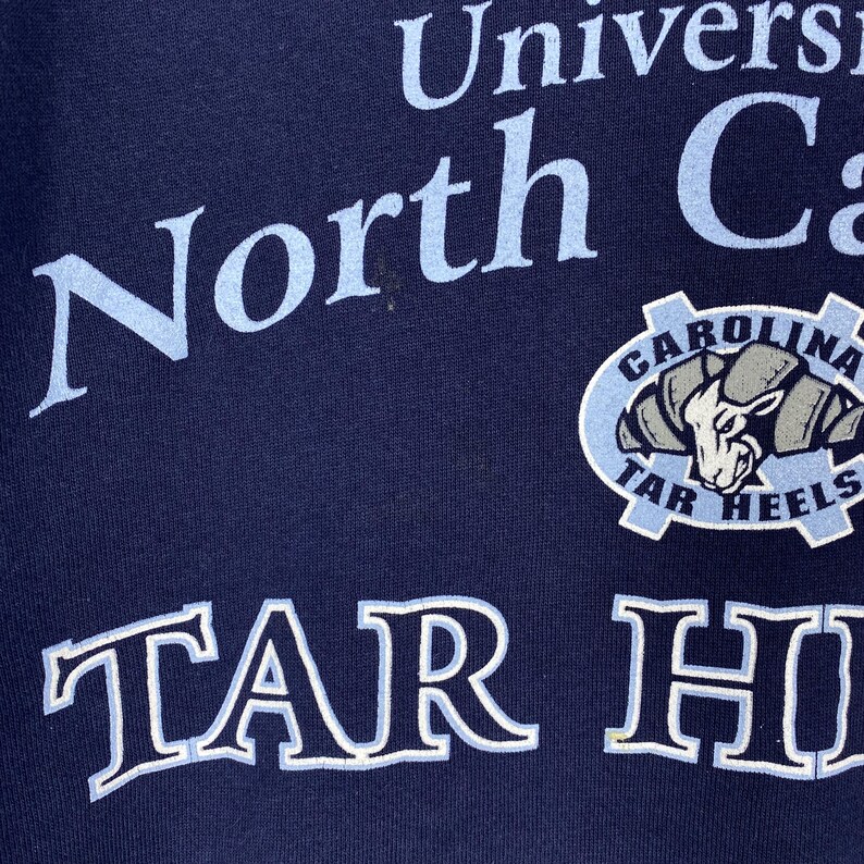 May include: Navy blue sweatshirt with the words "North Carolina" in light blue, and a "Tar Heels" logo. The logo features a ram's head in a light blue and grey colour scheme. The words "TAR HEELS" are also visible.
