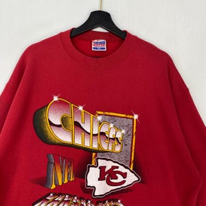 May include: A red crewneck sweatshirt featuring a graphic print with the text CHIEFS in a stylized font, the NFL logo, and the Kansas City Chiefs arrowhead emblem. The design includes sparkling star accents and a textured rectangular border.