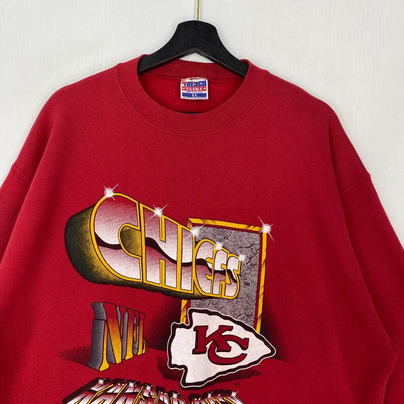 May include: A red crewneck sweatshirt featuring a graphic print with the text CHIEFS in a stylized font, the NFL logo, and the Kansas City Chiefs arrowhead emblem. The design includes sparkling star accents and a textured rectangular border.