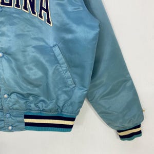 May include: Light blue satin jacket with the word "INA" in white and navy blue lettering. The jacket has a snap closure, a front pocket, and a striped cuff in navy, cream, and teal. The jacket is a vintage style.