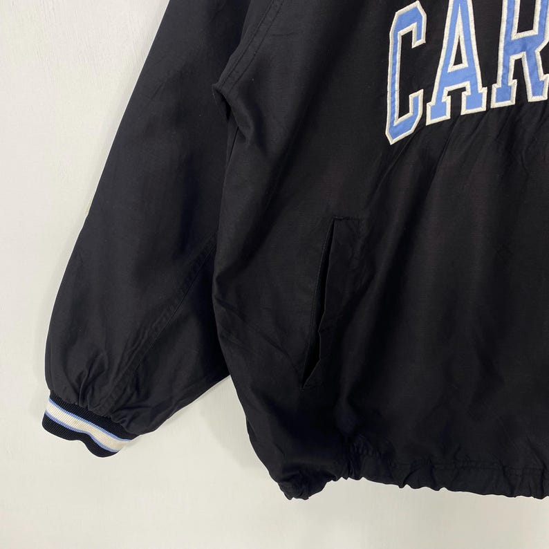 May include: Black jacket with the word "CAR" in light blue and white lettering across the chest. The jacket has a pocket on the front and a striped cuff on the sleeve. The jacket is made of a lightweight material.
