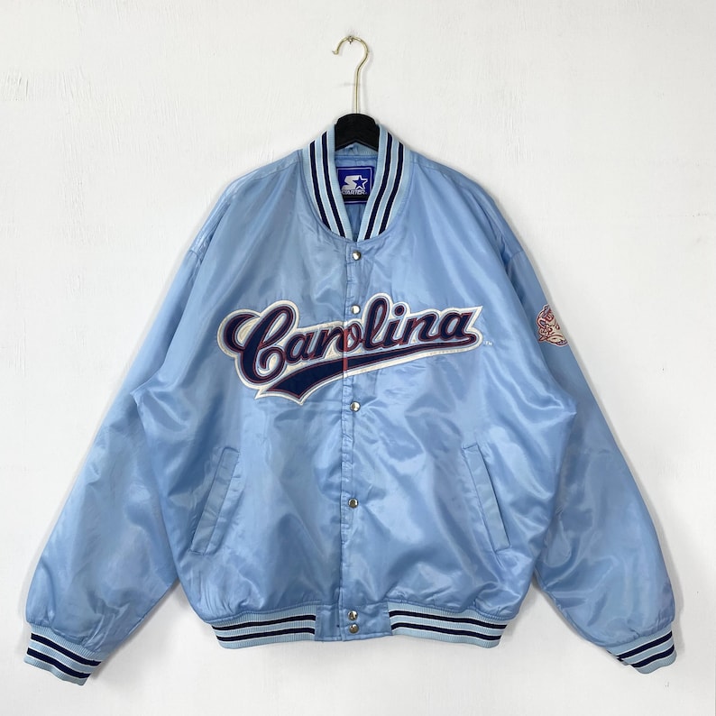 Puede incluir: Chaqueta de sat&eacute;n azul claro con la palabra "Carolina" en cursiva en el pecho. La chaqueta tiene una parte delantera abotonada, dos bolsillos y detalles a rayas en el cuello, los pu&ntilde;os y el dobladillo. Un peque&ntilde;o parche con el logo est&aacute; en la manga derecha.