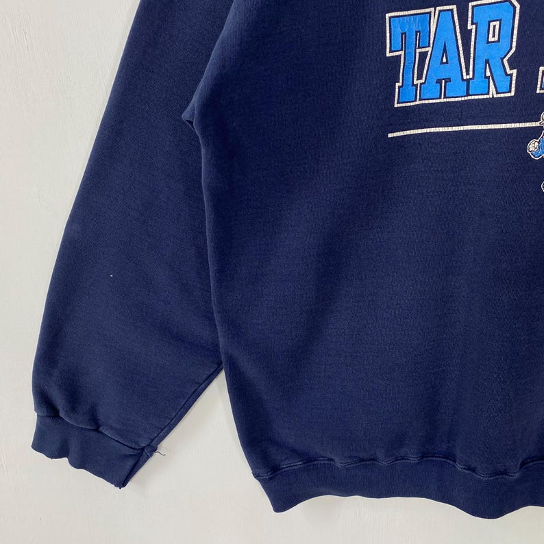 May include: Navy blue sweatshirt with the word "TAR" in white block letters. The sweatshirt has a crew neck and long sleeves. The image is a close-up of the upper portion of the sweatshirt.
