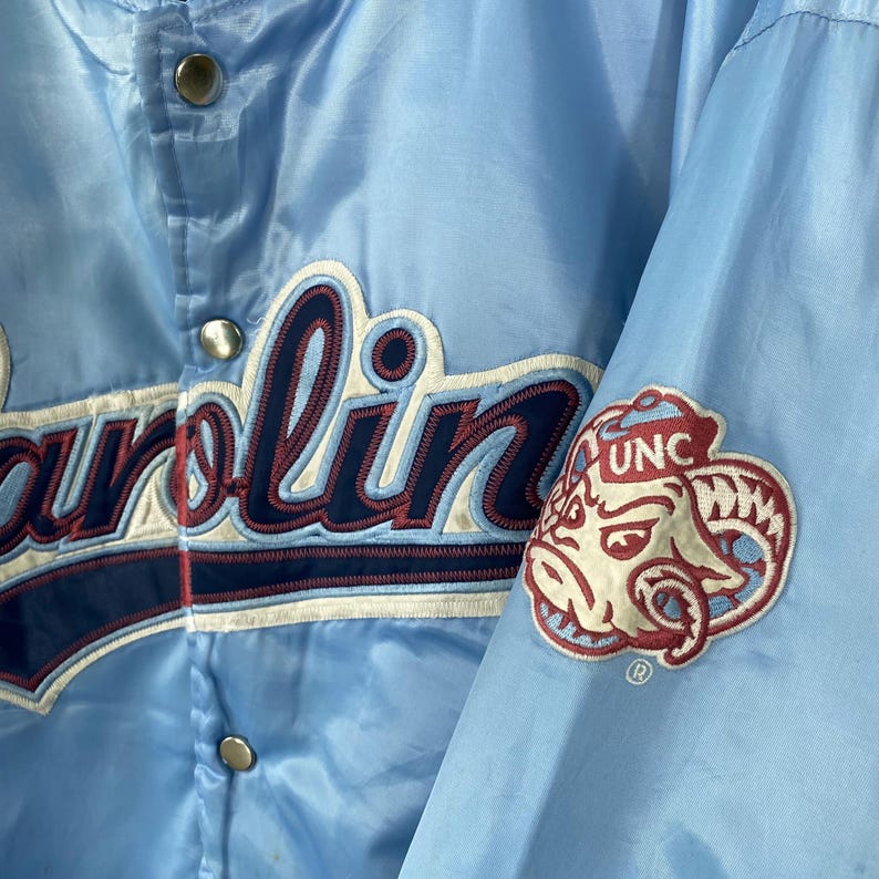 Puede incluir: Chaqueta de sat&eacute;n azul claro con la palabra "Carolina" bordada en blanco y azul marino. Un parche con un carnero y las letras "UNC" est&aacute; en la manga. La chaqueta tiene cierres de presi&oacute;n plateados.