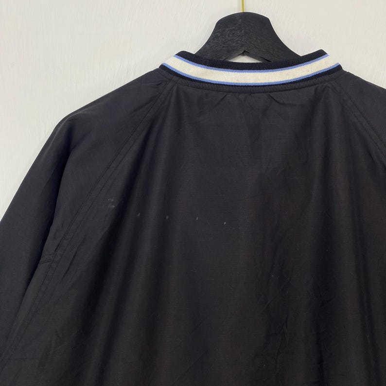 May include: Black jacket with a white, light blue, and navy blue striped collar. The jacket is hanging on a black hanger. The jacket appears to be made of a lightweight material.