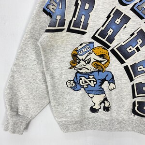 May include: A light grey sweatshirt featuring a graphic of a ram mascot wearing a blue sweater with the letters NC and a blue cap with UNC. The design includes large, bold, dark blue and light blue lettering that curves across the upper portion.
