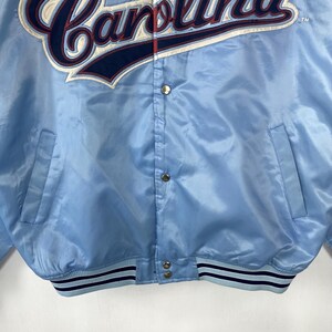 Puede incluir: Chaqueta de sat&eacute;n azul claro con la palabra "Carolina" bordada en blanco y azul marino en el pecho. La chaqueta tiene cierre de botones a presi&oacute;n, dos bolsillos delanteros y un dobladillo y pu&ntilde;os a rayas. Una chaqueta vintage de tem&aacute;tica deportiva.