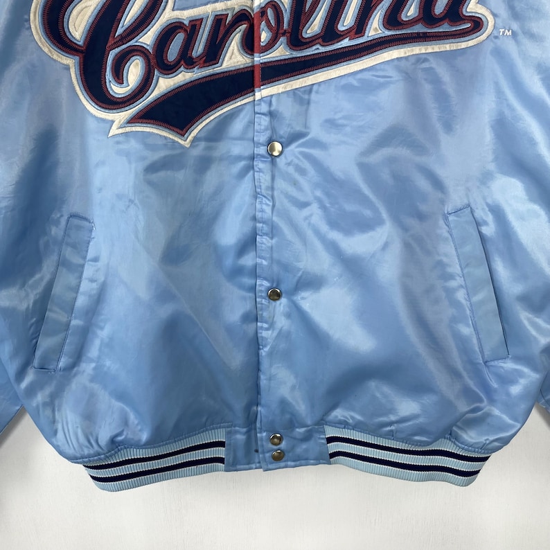 Puede incluir: Chaqueta de sat&eacute;n azul claro con la palabra "Carolina" bordada en blanco y azul marino en el pecho. La chaqueta tiene cierre de botones a presi&oacute;n, dos bolsillos delanteros y un dobladillo y pu&ntilde;os a rayas. Una chaqueta vintage de tem&aacute;tica deportiva.