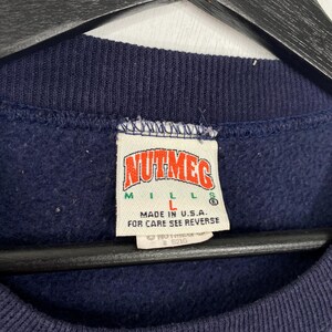 K&ouml;nnte beinhalten: Nahaufnahme eines marineblauen Sweatshirt-Etiketts. Das Etikett tr&auml;gt den Schriftzug "NUTMEG MILLS" in roter und wei&szlig;er Schrift. Darunter steht "MADE IN U.S.A." und "FOR CARE SEE REVERSE". Das Etikett ist wei&szlig; mit blauem Rand.