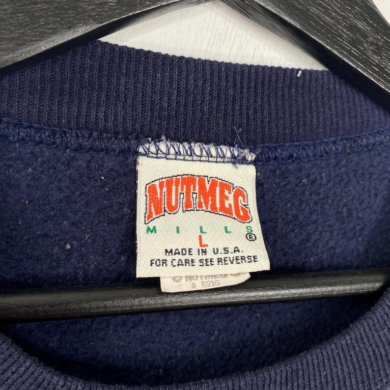 K&ouml;nnte beinhalten: Nahaufnahme eines marineblauen Sweatshirt-Etiketts. Das Etikett tr&auml;gt den Schriftzug "NUTMEG MILLS" in roter und wei&szlig;er Schrift. Darunter steht "MADE IN U.S.A." und "FOR CARE SEE REVERSE". Das Etikett ist wei&szlig; mit blauem Rand.