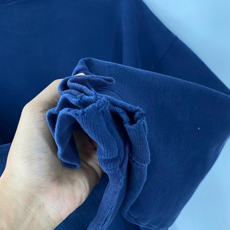May include: Close-up of a navy blue sweatshirt sleeve. The ribbed cuff is bunched up, revealing the texture of the fabric. The background is a lighter shade of blue.
