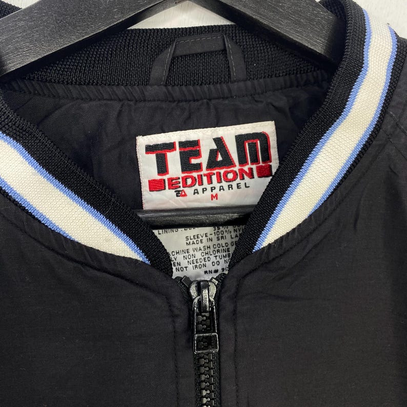 May include: Black bomber jacket with a full zipper and a white, light blue, and black striped collar. The jacket has the words "TEAM EDITION APPAREL" on a tag.