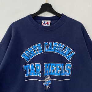 May include: Navy blue sweatshirt with the words "North Carolina Tar Heels" in white and light blue. The sweatshirt features a Tar Heels mascot graphic and a Logo Athletic tag.