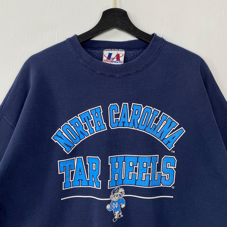 May include: Navy blue sweatshirt with the words "North Carolina Tar Heels" in white and light blue. The sweatshirt features a Tar Heels mascot graphic and a Logo Athletic tag.