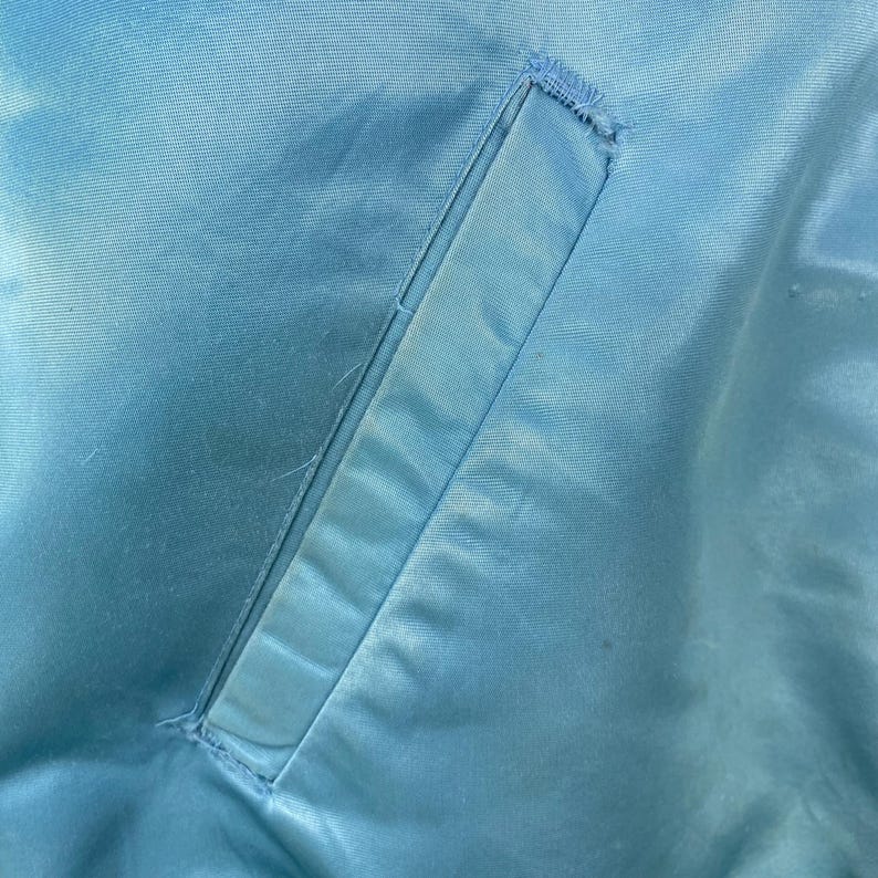 May include: Close-up of a light blue satin jacket. The image highlights a rectangular pocket with a stitched border. The fabric has a subtle sheen, and the pocket is slightly darker than the surrounding material. The jacket appears to be vintage.