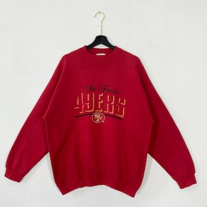 May include: A red sweatshirt featuring the embroidered text "San Francisco 49ers" in gold and black. The sweatshirt has a crew neck and long sleeves. The 49ers logo is embroidered below the text.