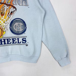 May include: A light blue long-sleeve sweatshirt featuring a graphic of a basketball hoop with a flaming basketball. The design includes the text CAROLINA and HEELS, along with a circular logo containing the letters NC in a dark blue color.