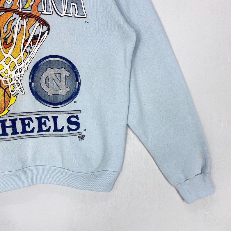 May include: A light blue long-sleeve sweatshirt featuring a graphic of a basketball hoop with a flaming basketball. The design includes the text CAROLINA and HEELS, along with a circular logo containing the letters NC in a dark blue color.