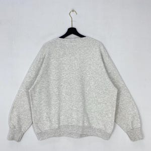 May include: A light grey heathered crewneck sweatshirt with long sleeves and ribbed cuffs at the wrists and waist. The garment is displayed on a black plastic hanger against a plain, light-coloured background, showing the back side of the piece.