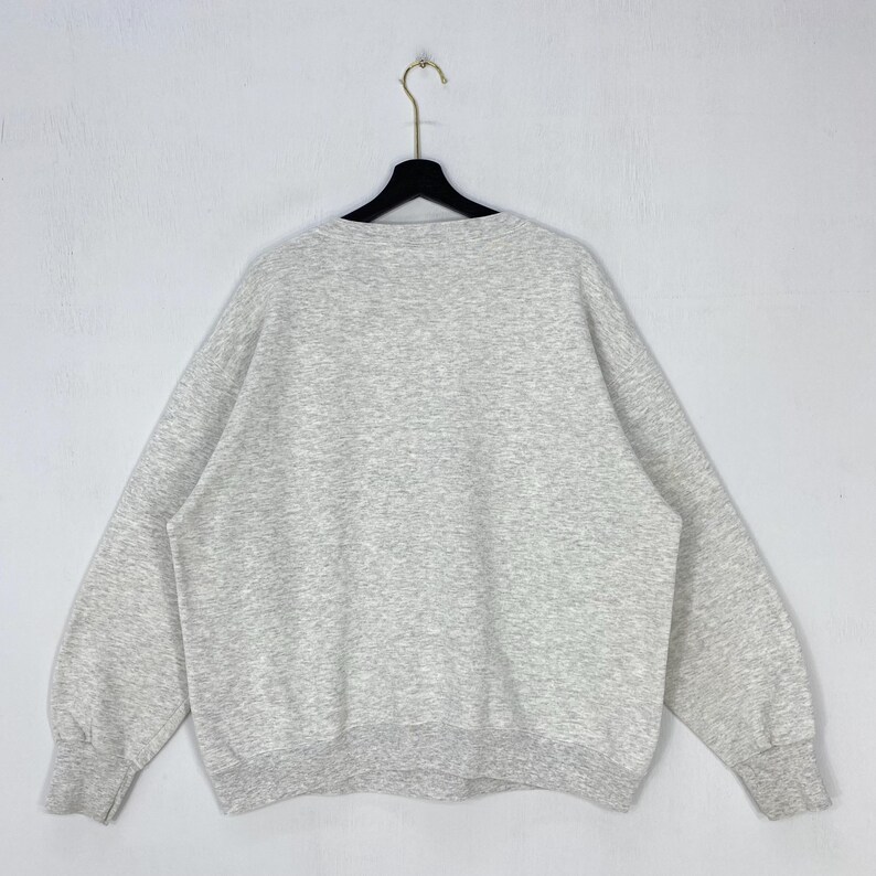 May include: A light grey heathered crewneck sweatshirt with long sleeves and ribbed cuffs at the wrists and waist. The garment is displayed on a black plastic hanger against a plain, light-coloured background, showing the back side of the piece.