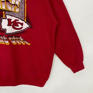 May include: A red long-sleeve sweatshirt featuring a graphic print of the Kansas City Chiefs logo and text. The design includes a white arrowhead shape with a red KC emblem, along with stylized lettering that reads Kansas City in yellow and white.