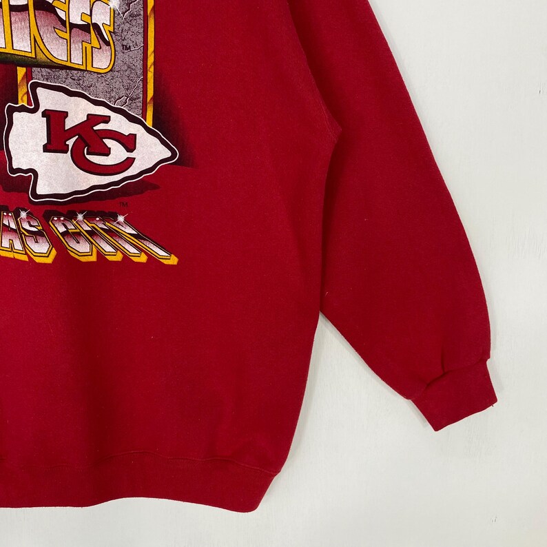 May include: A red long-sleeve sweatshirt featuring a graphic print of the Kansas City Chiefs logo and text. The design includes a white arrowhead shape with a red KC emblem, along with stylized lettering that reads Kansas City in yellow and white.