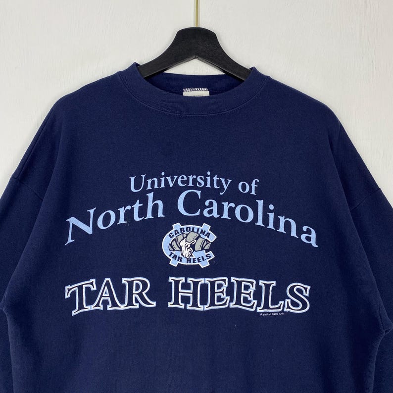 May include: A navy blue sweatshirt with the words "University of North Carolina" in light blue lettering. Below the text is a logo with the words "Tar Heels". The sweatshirt is suspended from a black hanger.
