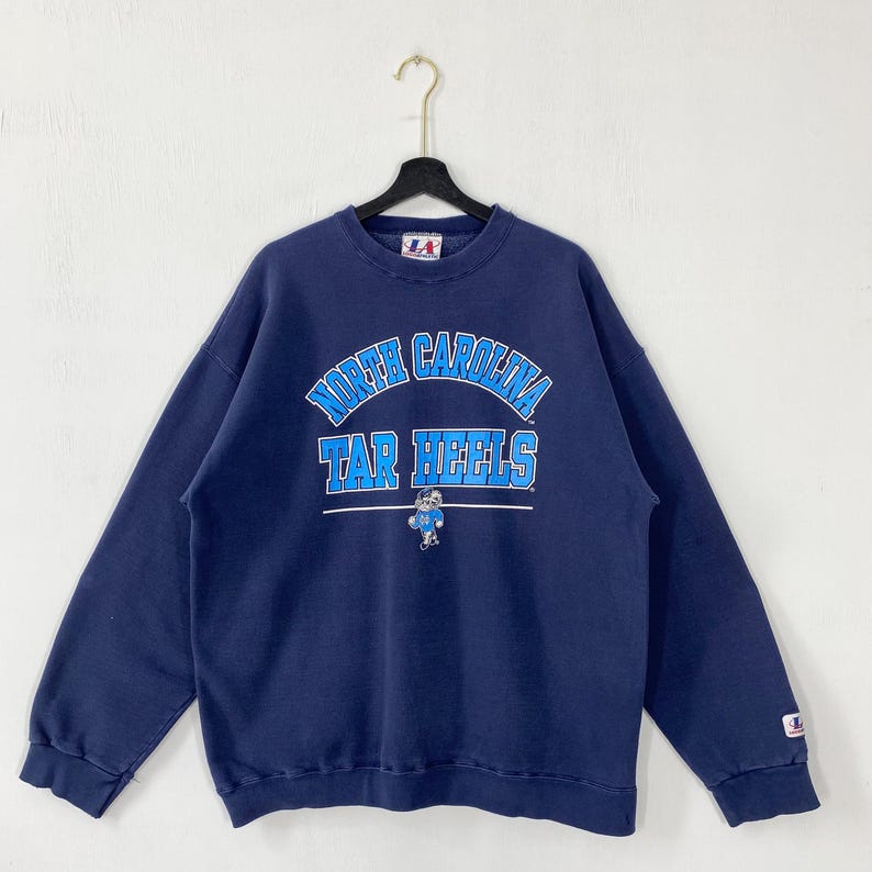 May include: Navy blue sweatshirt with the words "North Carolina Tar Heels" in light blue lettering. A cartoon Tar Heel mascot is below the text. The sweatshirt is hanging on a black hanger.