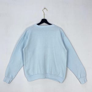 May include: A light blue long-sleeve crewneck sweatshirt featuring a solid color design, a ribbed collar, ribbed cuffs, and a ribbed hem. The garment is displayed hanging on a black plastic hanger against a plain, neutral-colored wall.