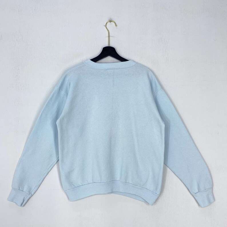 May include: A light blue long-sleeve crewneck sweatshirt featuring a solid color design, a ribbed collar, ribbed cuffs, and a ribbed hem. The garment is displayed hanging on a black plastic hanger against a plain, neutral-colored wall.