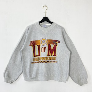 May include: A heather gray sweatshirt with the text "U of M Gophers" in maroon and gold. The sweatshirt has long sleeves and a crew neck. The graphic includes a maroon "M" on each side of the text and a gold emblem.