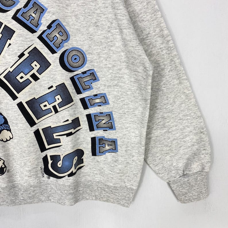 May include: A light grey long-sleeve sweatshirt featuring the text CAROLINA HEELS in large, bold, blue and black block lettering. The design includes a partial graphic of a character on the left side and the brand name DECOTEX printed below.
