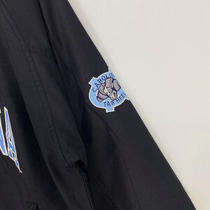 May include: Black jacket with a light blue and white logo patch on the sleeve. The patch features a ram and the words "CAROLINA" and "TAR HEELS". The jacket has a zipper and a simple design.