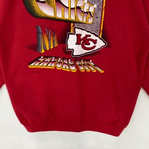 May include: A red sweatshirt featuring a graphic print with the text CHIEFS, NFL, and KANSAS CITY. The design includes a white arrowhead logo with red KC lettering. The graphic elements are rendered in a bold, dimensional style with yellow and black accents.