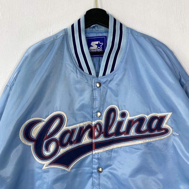 Puede incluir: Chaqueta de sat&eacute;n azul claro con la palabra "Carolina" en una fuente cursiva en la parte delantera. La chaqueta tiene un cuello a rayas y el logotipo de Starter. La chaqueta est&aacute; abotonada y colgada de una percha negra.